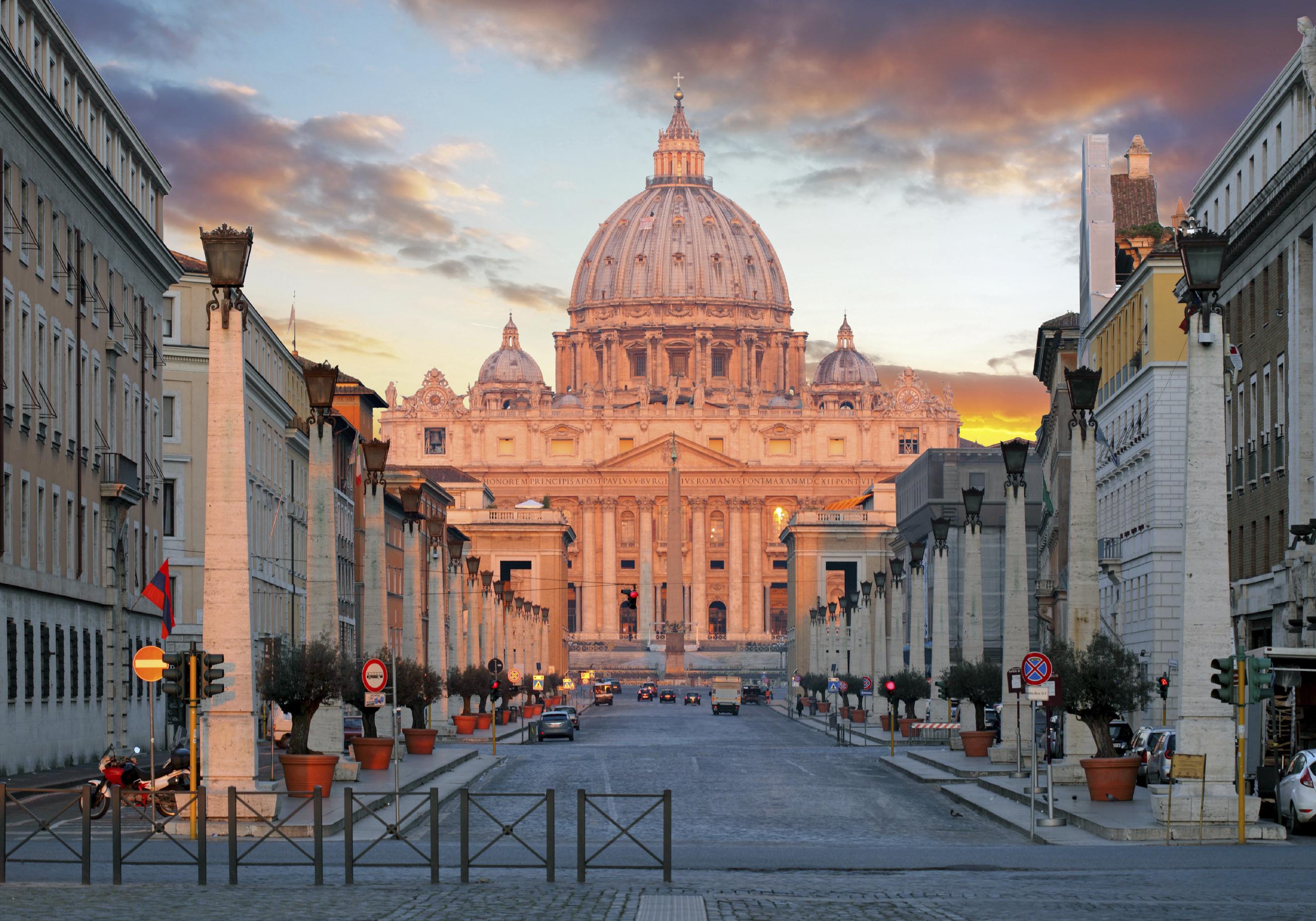 St. Peter's Basilica in Rome, illuminated by the warm light of sunset, towers at the end of a wide street lined with buildings. Dramatic clouds adorn the sky.