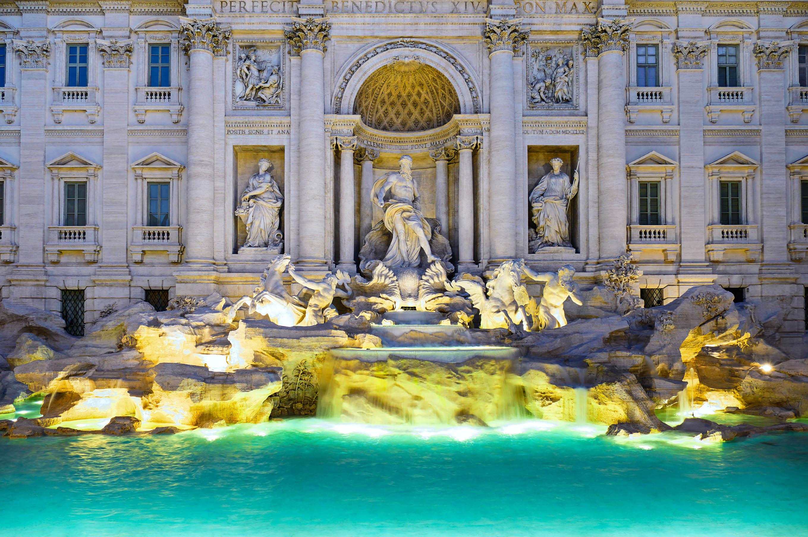 The Trevi Fountain in Rome at night. At the center of the fountain stands a large sculpture, surrounded by artistic figures. The water glows turquoise, and the facade is brightly illuminated.