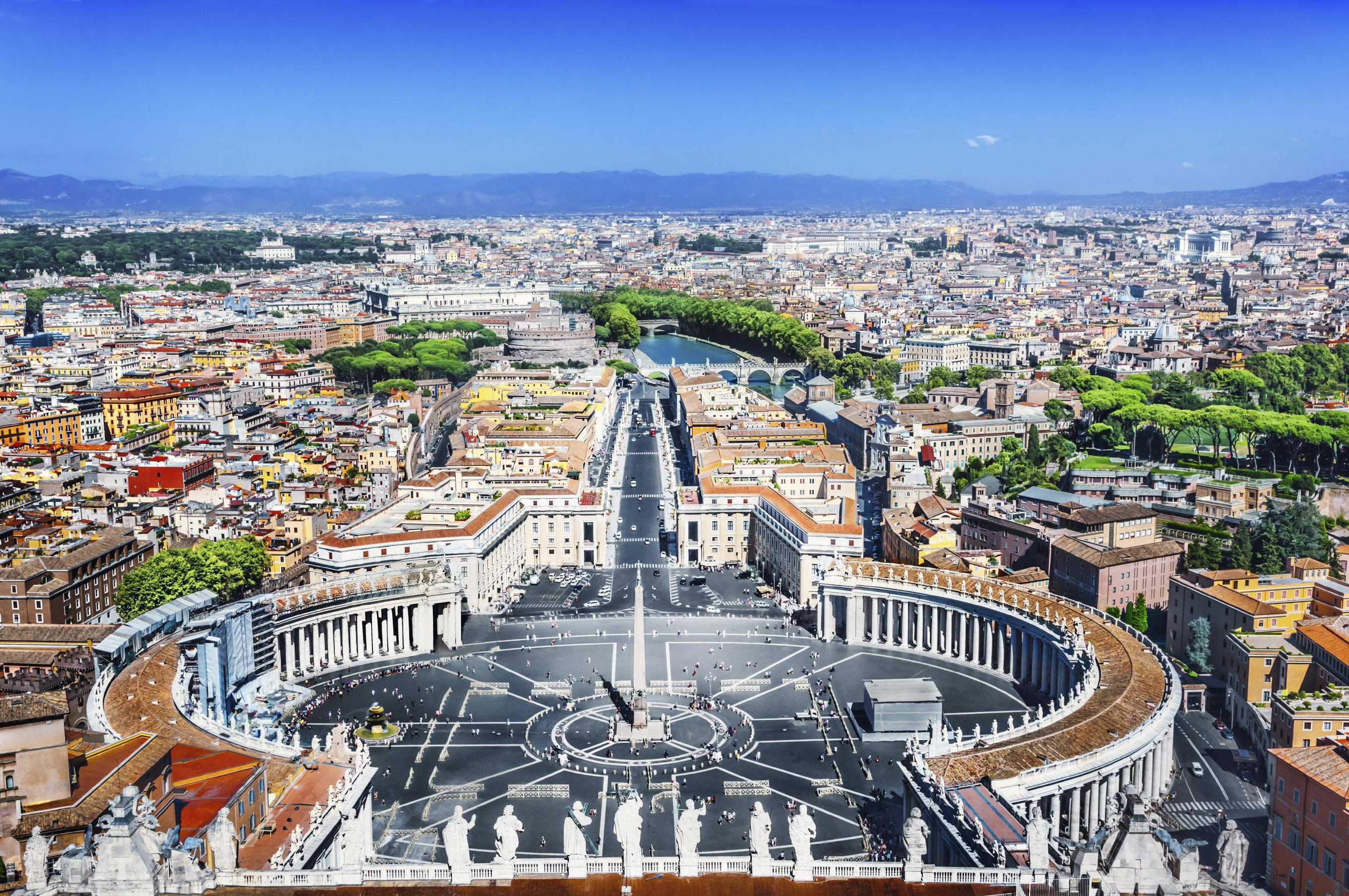 View of St. Peter's Square in Rome with its oval colonnade and central obelisk. The city extends in the background with a green park and blue sky above.
