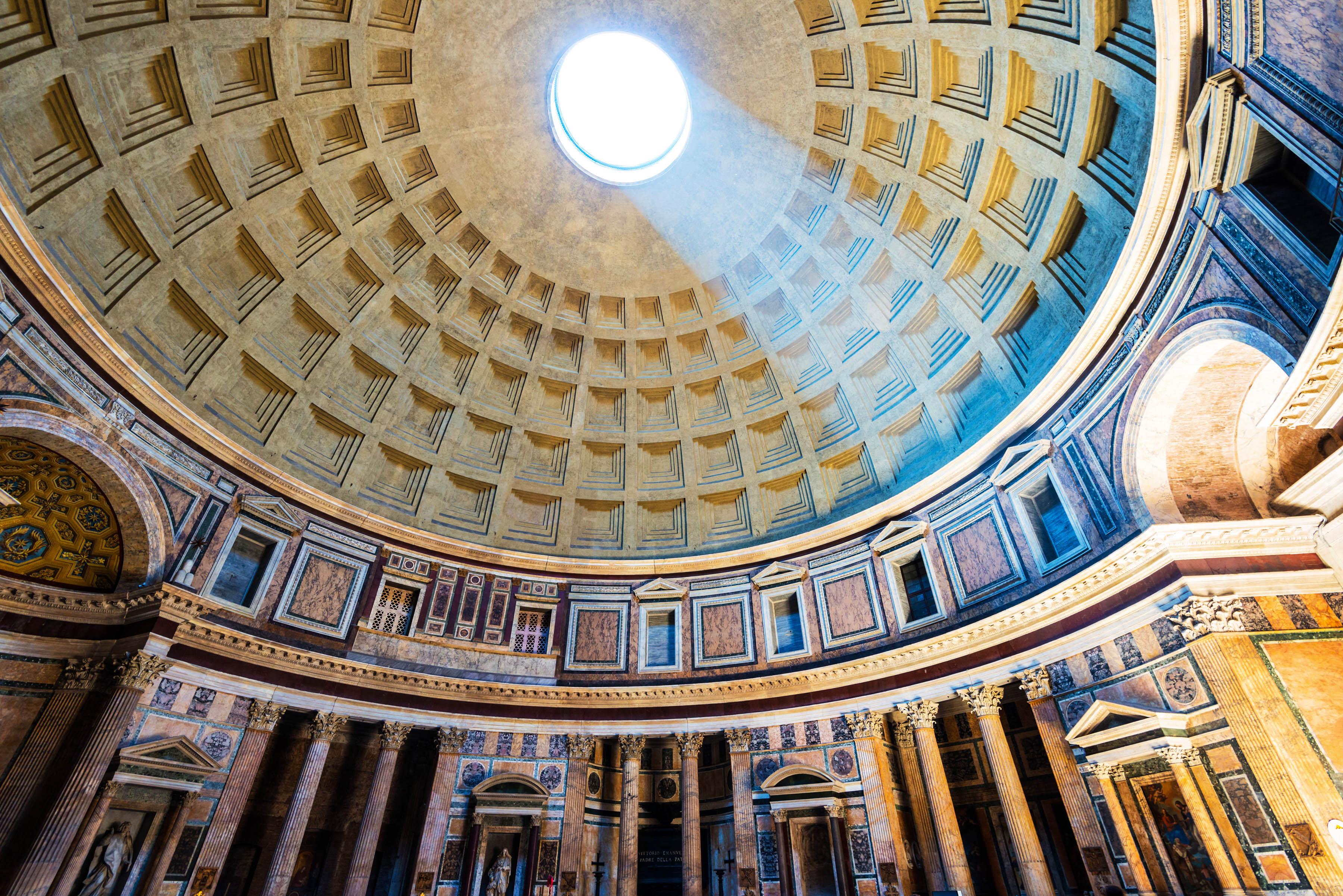 Beams of light streaming through the circular opening in the dome of the Pantheon in Rome. Marble columns and intricate decorations adorn the rounded inner wall, creating a majestic ambiance.