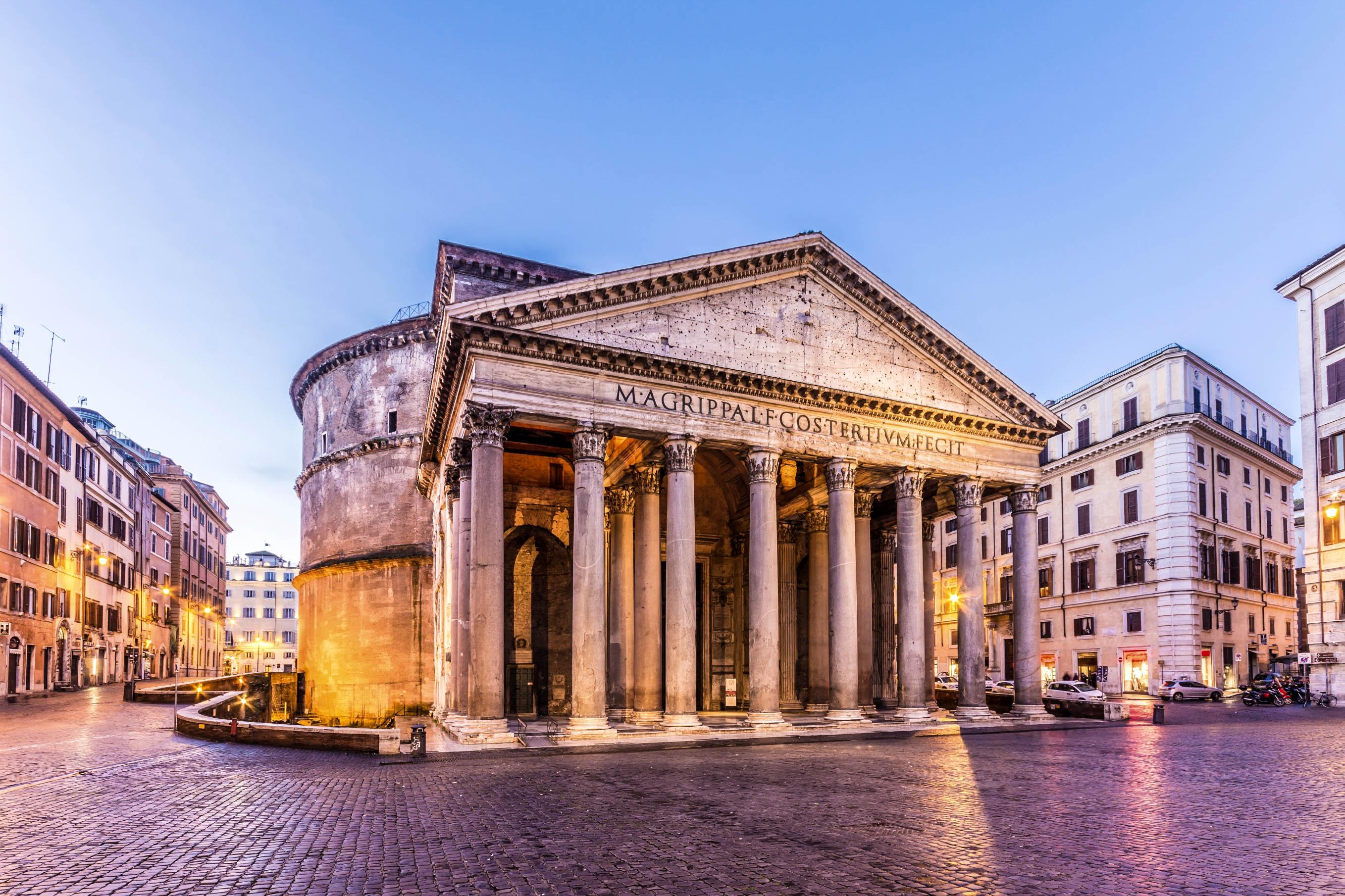 The Pantheon in Rome at dusk, taken from the front. Massive columns support a pediment; behind it, the round dome arches. Surrounded by buildings, cobblestones in the foreground.