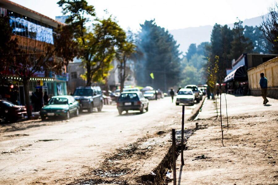 Dusty street with cars and pedestrians, buildings and trees on the left. On the right, an unpaved sidewalk with small trees. Mountains and more trees visible in the background. Bright, sunny atmosphere.