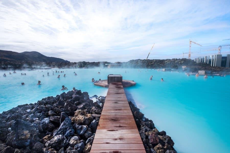 A wooden bridge extends from the middle of the lower edge of the image to the center. The wooden bridge leads into the brilliantly blue water of the Blue Lagoon, where many people are bathing. Steam rises, and mountains are visible in the background.
