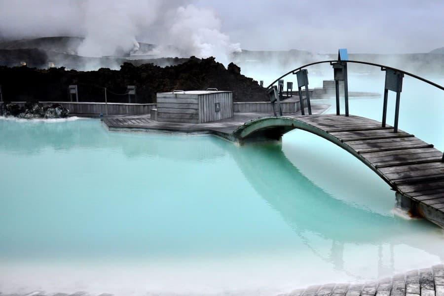 On the right edge of the image, there is a wooden bridge that arches over the water of the Blue Lagoon. The water is light blue, almost white in some spots. In the background, mountains are visible but obscured by dense fog.