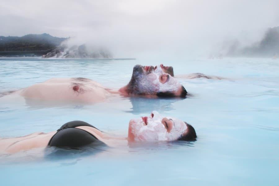 In the foreground of the image, a woman and a man are seen, half of their upper bodies in the picture. They are lying on their backs in the water of the Blue Lagoon - only their upper bodies and faces are visible. They also have white masks on their faces.