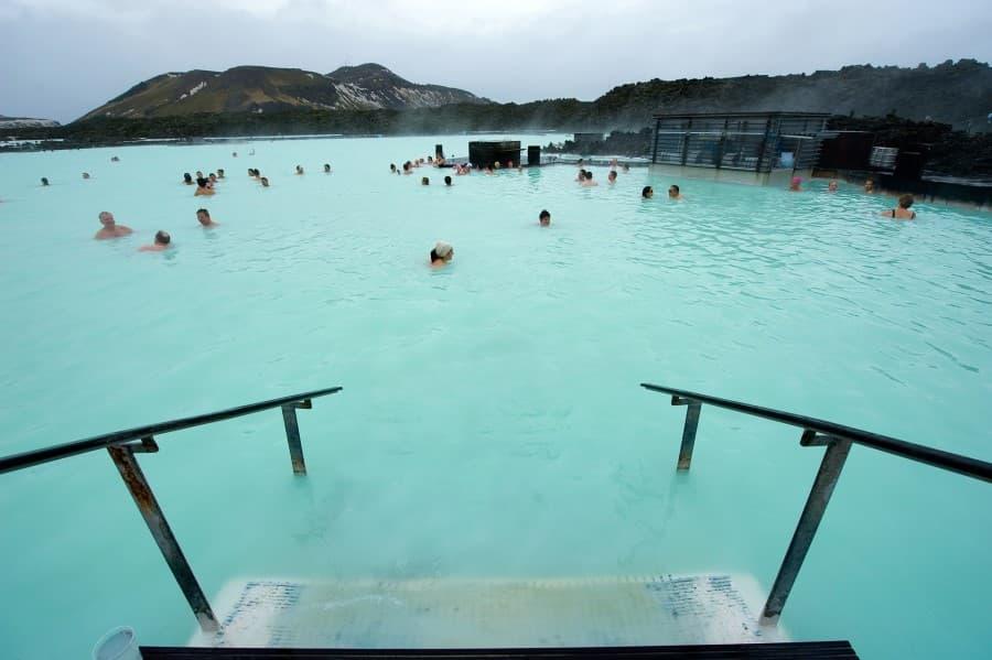 Almost the entire picture consists of the light blue water of the Blue Lagoon in Iceland, with many visitors' heads protruding from it. In the foreground, a staircase can be seen leading into the water. In the background, mountains rise into the sky.