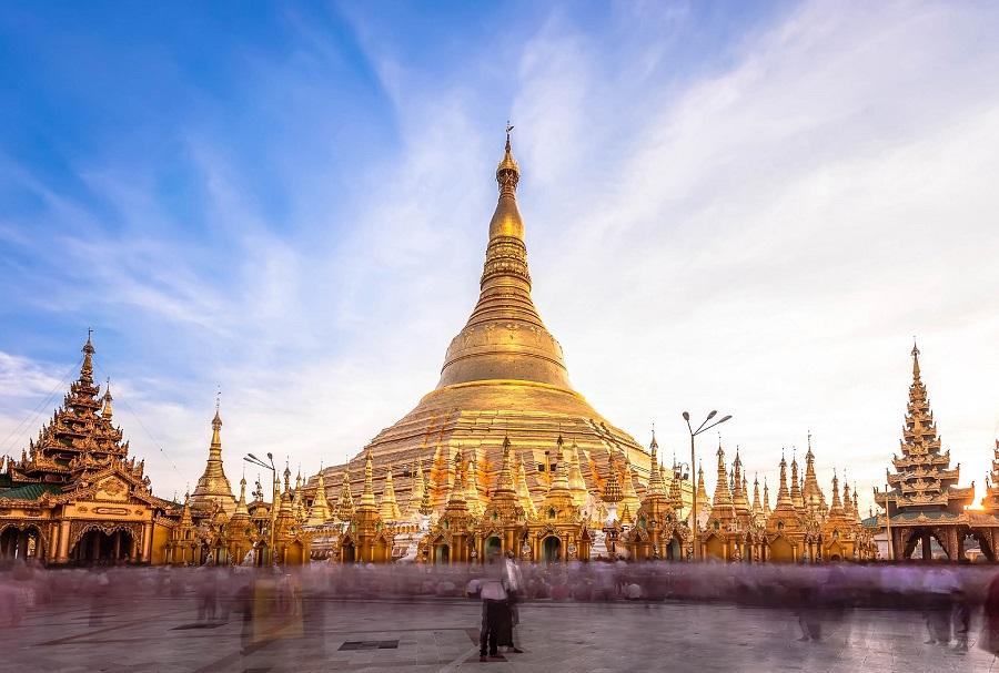 Golden Pagoda with a pointed tower in the center, flanked by smaller towers and decorative buildings. People are moving in the foreground while the sky shines blue and white.