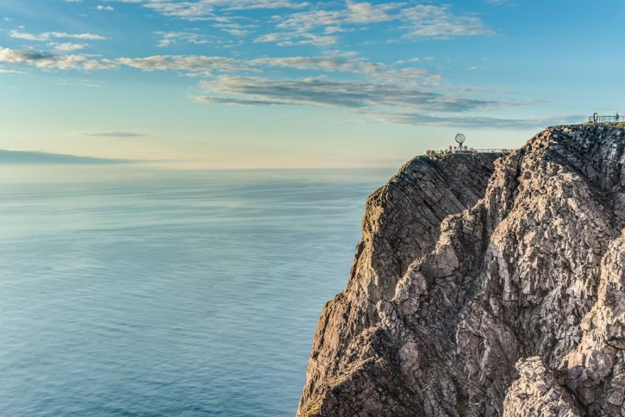 On the right side of the picture lies the cliff of the North Cape, topped with the Globe statue. On the left is the sea, above the blue sky with a few clouds.