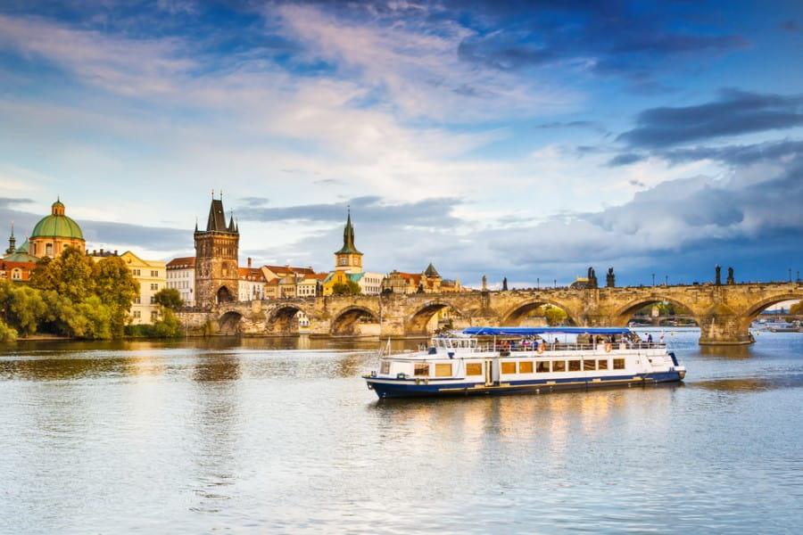 Urban skyline with historical buildings on both sides of a stone bridge. In the foreground, a river cruise ship passes by. Perfect for October cruises with autumnal ambiance.
