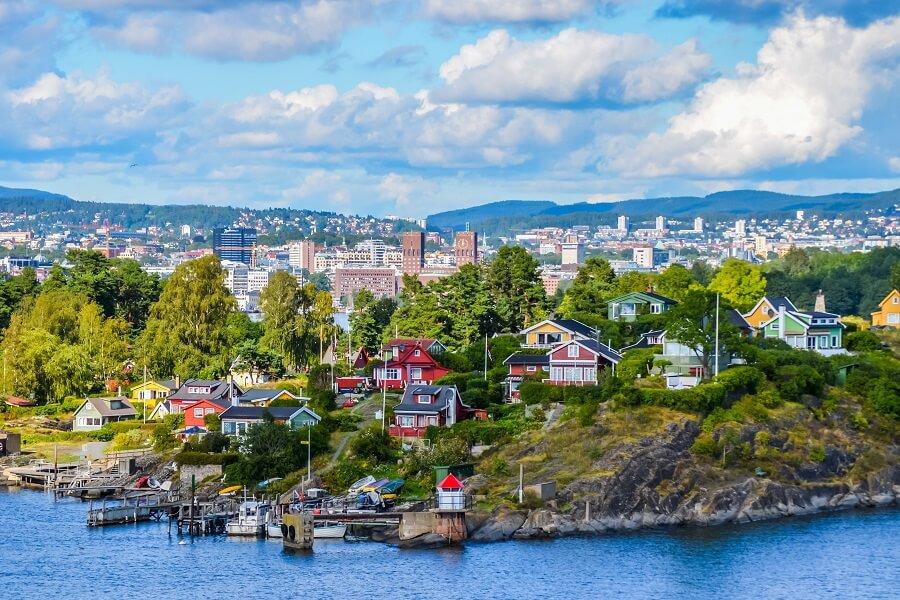 View of the Oslofjord. At the bottom of the image, the river flows, above is the coast with nature and houses. In the background, you can see Oslo and mountains. The sky is blue but cloudy.