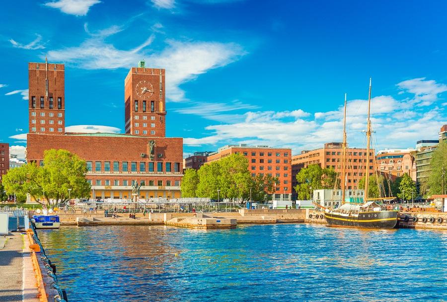 Photo of the Oslo City Hall, made of red stone. In the foreground is water, reflecting the building. The sky is blue.