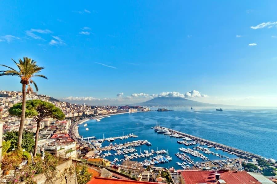 Aerial view of a Neapolitan harbor, stretching to the left of the image. To the right is the sea. In the background, Mount Vesuvius rises.