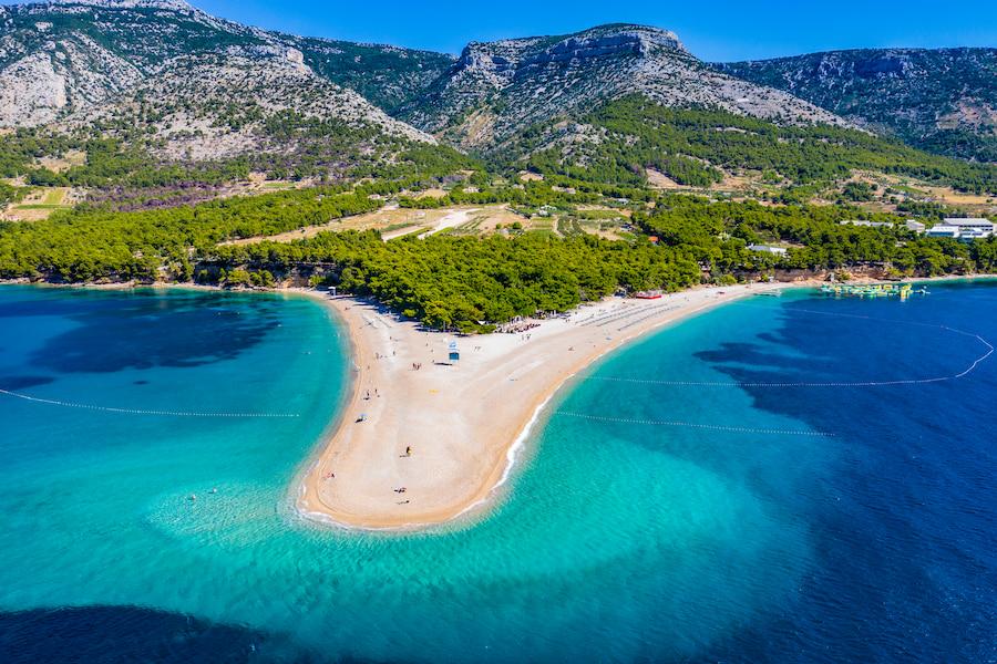 The Zlatni Rat beach in Bol on the island of Brač. Surrounded by turquoise water, the golden horn extends far into the sea. In the background, you see mountains and green landscapes.