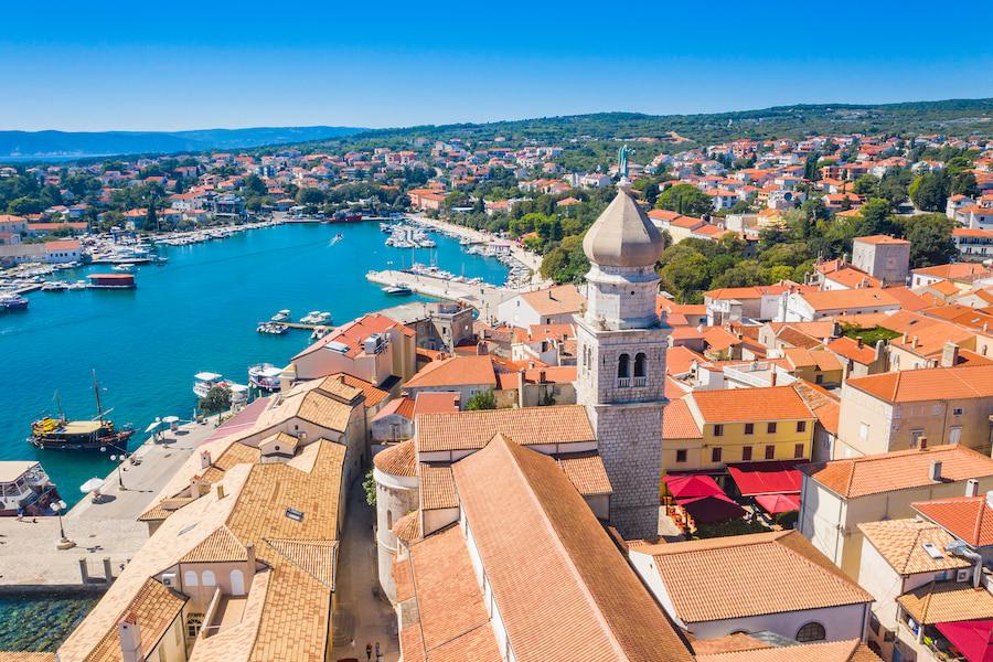 Aerial view of the picturesque old town of Krk in Croatia. In the foreground, numerous buildings and a church tower can be seen. To the left, there is the idyllic harbor of the old town. In the background, scattered buildings and the green landscape of the beautiful island with the sea behind it can be seen.