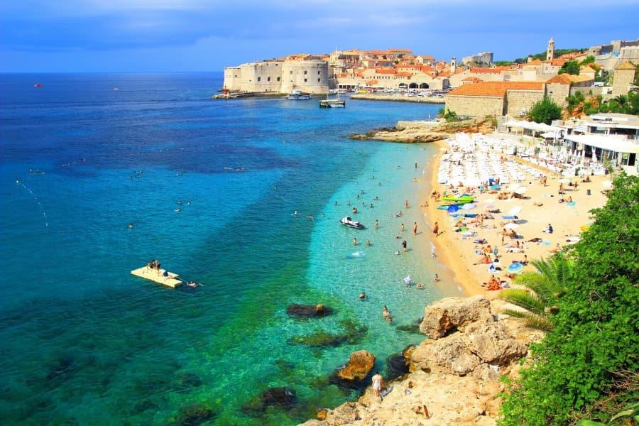 The image on the left shows the sea of the Adriatic coast, which brightens from deep blue to turquoise as it approaches the shore. In the background on the right, the city with many small houses can be seen. In the foreground, a well-populated beach is visible, furnished with white lounge chairs and a beach bar.