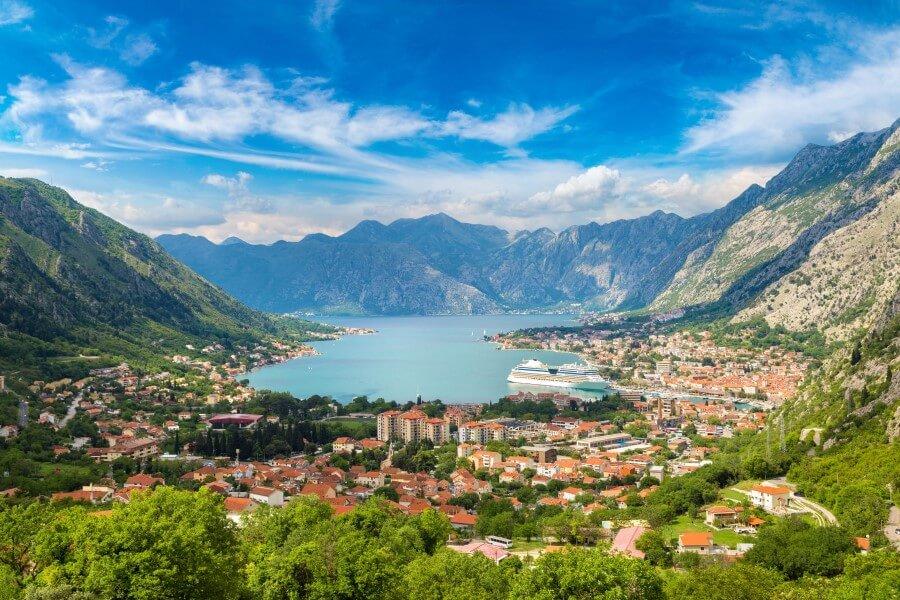 View of the Bay of Kotor, surrounded by green mountains. A city with red roofs and white buildings lies below. The blue sky is partly cloudy. The sea extends in the middle.