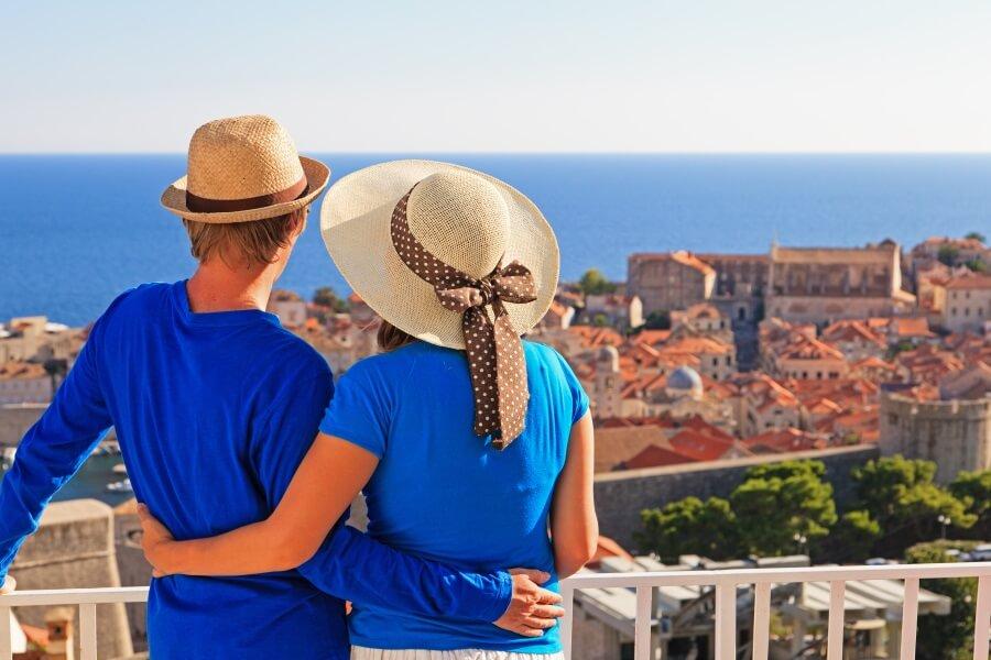 A couple in blue shirts stands on a balcony, embracing and looking over Dubrovnik with the blue sea on the horizon. Both are wearing straw hats, the one on the right has a ribbon.