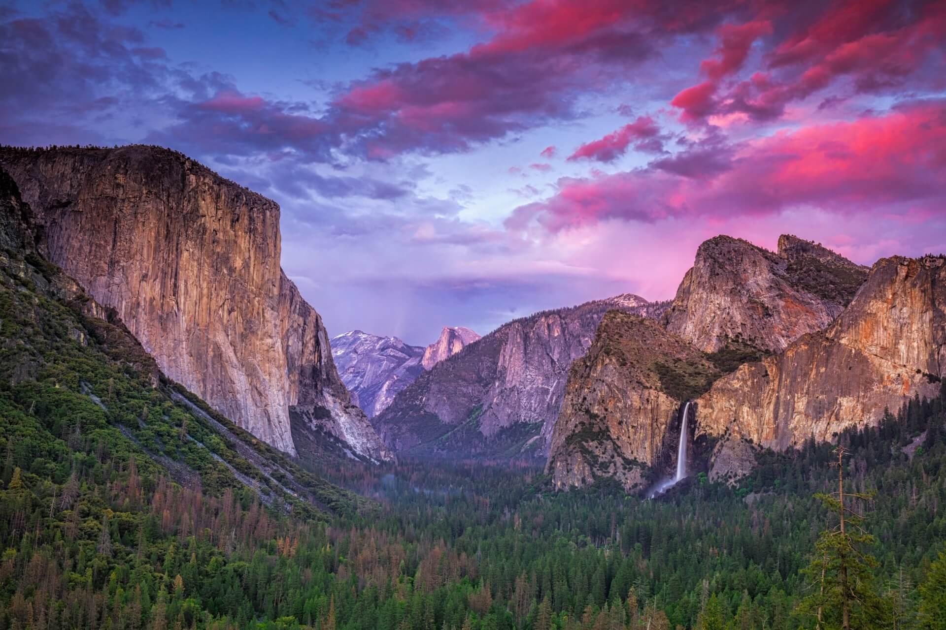 View of Yosemite Valley: on the left, the striking rock wall El Capitan; in the middle, distant mountains; on the right, waterfall against a rocky backdrop. The sky is colorful, predominantly purple and pink, beneath a dense forest.
