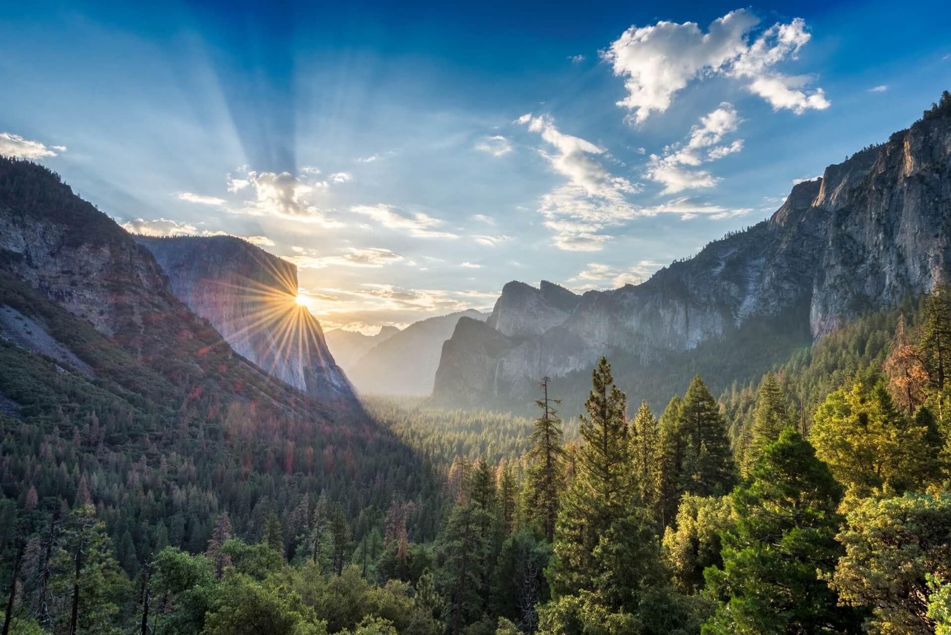 The sun sets behind the mountain backdrop, shining through a valley surrounded by dense forests. On the left, high cliffs; on the right, wooded hills. The sky is blue with light clouds.