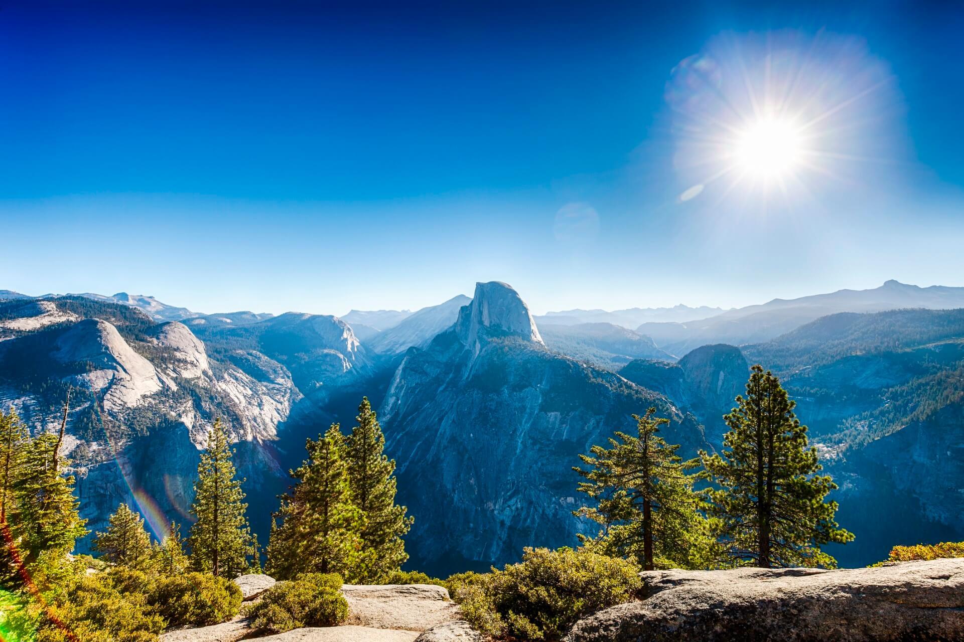 View from Glacier Point over Yosemite Valley with the iconic Half Dome at the center, surrounded by forested mountains. In the foreground, green trees and shrubs, above, a shining sun in a clear sky.
