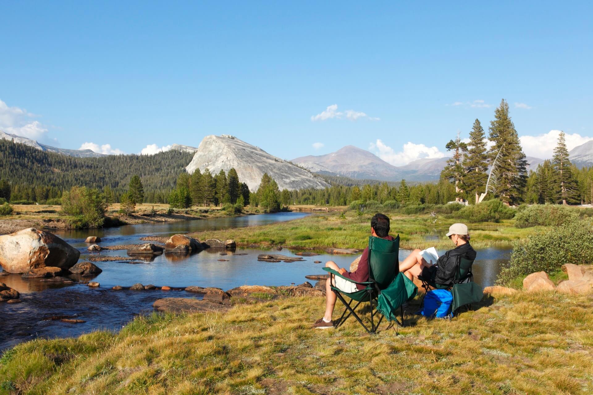Two people sit in camping chairs by the shore of a clear river, surrounded by meadows and trees. In the background, impressive mountain peaks of Yosemite National Park rise under a blue sky.