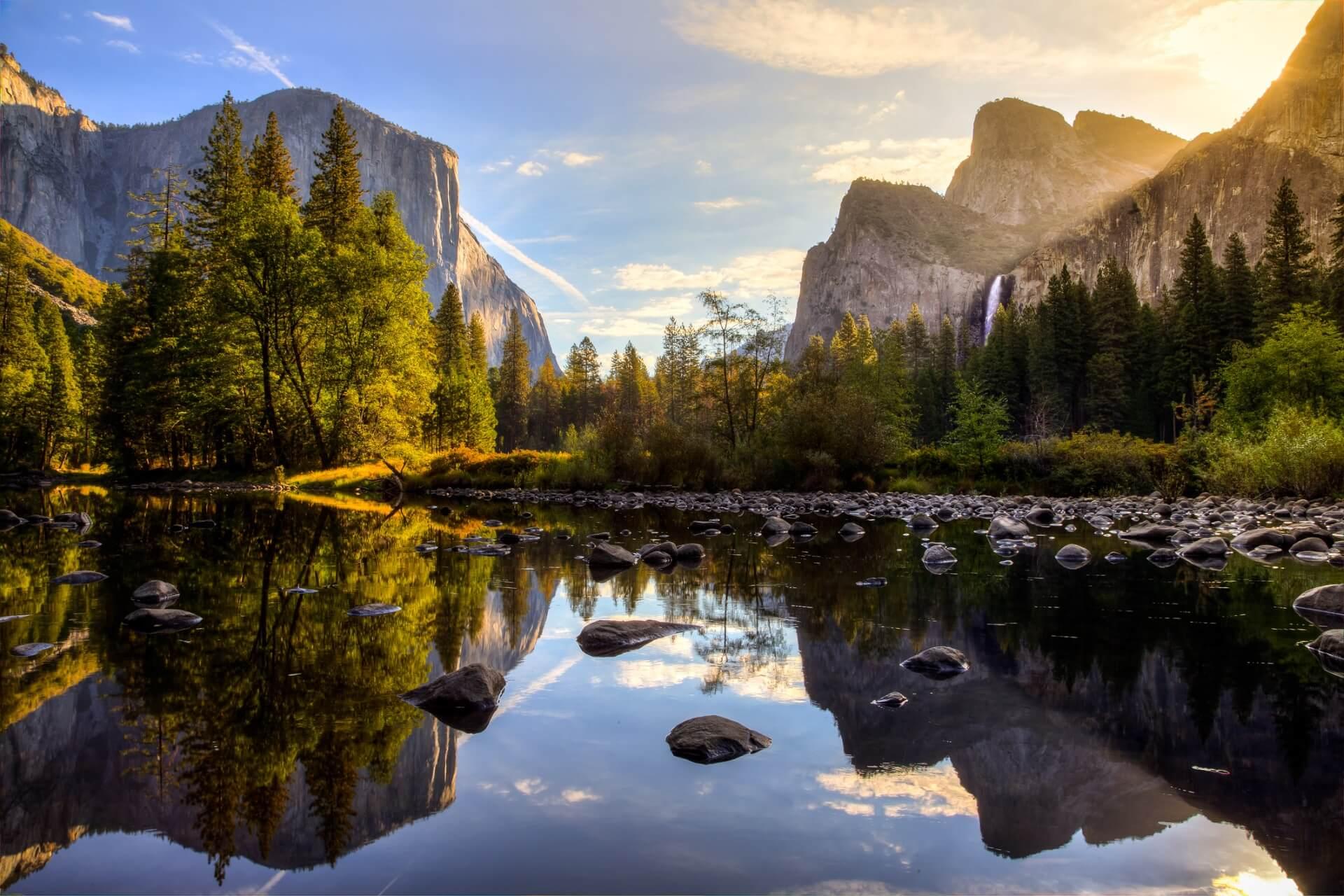 Sunrise in Yosemite Valley: On the left is a large, steep rock surrounded by green trees. On the right, a waterfall cascades down a wooded cliff. In the foreground, a calm river with stones.