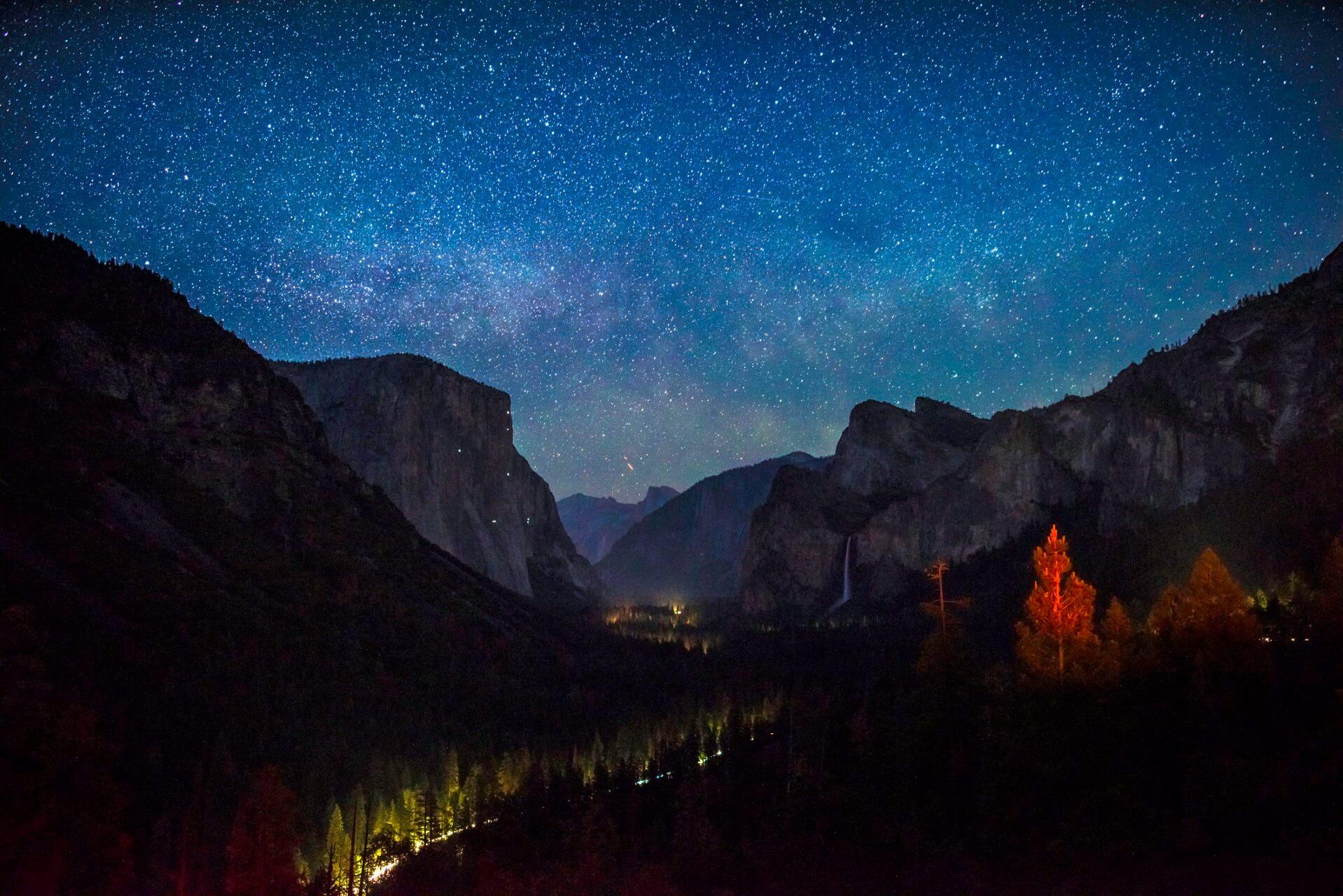 Starry night over Yosemite Valley with tall rocks, with the iconic El Capitan on the left and a waterfall on the right. Trees in the foreground glow golden.