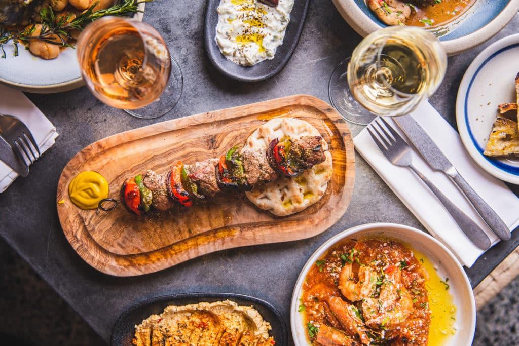 a table covered in greek food and wine, with a platter of souvlaki taking the place of pride in the middle