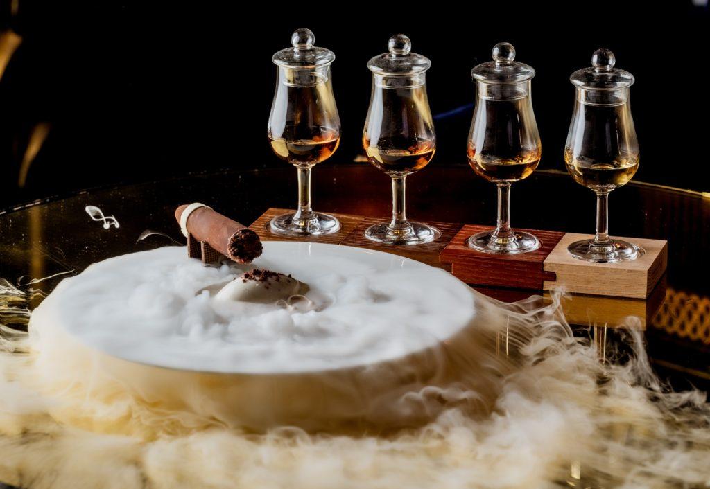 a chocolate cigar perched on a plate, with dry ice smoke billowing off the plate, with four whiskey samples behind